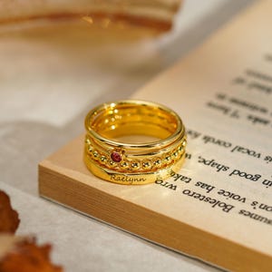 Könnte beinhalten: Ein Stapel goldfarbener Ringe, einer davon mit einem kleinen roten Edelstein und dem eingravierten Namen "Raelynn". Die Ringe liegen auf einem Buch.