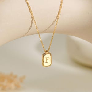 Könnte beinhalten: Eine goldfarbene Halskette mit einer feinen gedrehten Kette und einem rechteckigen Anhänger mit texturiertem Rand. In der Mitte des Anhängers befindet sich der Großbuchstabe F in einer Serifenschrift mit polierter, reflektierender Oberfläche.