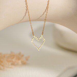 May include: A gold-toned necklace featuring a delicate chain and a pendant shaped like an open heart outline with flame-like edges. The pendant is suspended from the center of the chain, which has small, uniform oval links throughout its length.