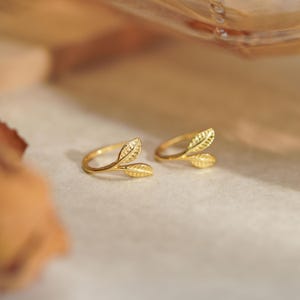 Op de afbeelding: Twee goudkleurige ringen in de vorm van bladeren. Elke ring heeft een delicaat ontwerp van twee bladeren, met ingewikkelde details op het oppervlak. De ringen staan tegen een zachte, neutrale achtergrond, wat hun elegante en natuurlijke esthetiek benadrukt.