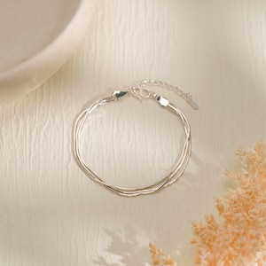 Könnte beinhalten: Ein mehrsträngiges silberfarbenes Armband aus dünnen, flexiblen Schlangenkettengliedern, die sich überlappen und einen Lagen-Look erzeugen. Das Armband verfügt über einen kleinen Karabinerverschluss und eine verstellbare Verlängerungskette.