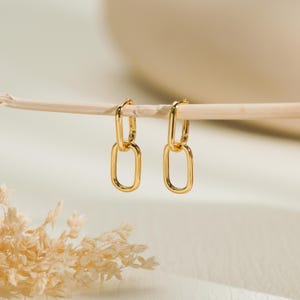 May include: A pair of gold-colored hoop earrings with a linked oval design. The earrings are suspended from a thin, light-colored wooden stick against a soft, neutral background. The earrings have a polished finish.