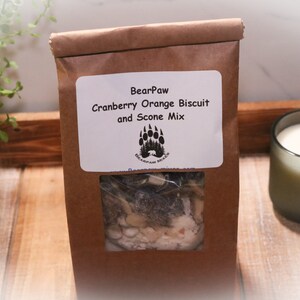 May include: A brown paper bag with a clear window showing a cranberry orange biscuit and scone mix. The bag has a label that says "BearPaw Cranberry Orange Biscuit and Scone Mix".