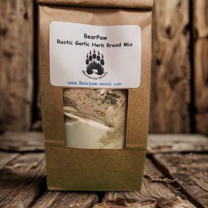 May include: A brown paper bag of BearPaw Rustic Garlic Herb Bread Mix. The label features a bear paw logo and the website address. A clear window reveals the bread mix inside. The bag is set against a rustic wooden background.