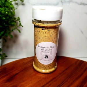 May include: A glass jar with a white lid filled with a yellow and green spice blend. The label reads "Bearpaw Mixes Beary Butter Steak Butter Mix" with a bear paw logo and the website address "www.bearpaw-mixes.com".