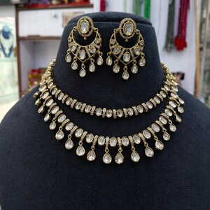 May include: A gold-toned necklace and earring set featuring clear oval-shaped stones. The necklace consists of two layered chains with dangling teardrop accents. The matching earrings are crescent-shaped with a central stone and hanging drops.