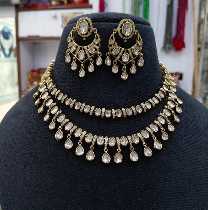 May include: A gold-toned necklace and earring set featuring clear oval-shaped stones. The necklace consists of two layered chains with dangling teardrop accents. The matching earrings are crescent-shaped with a central stone and hanging drops.