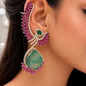 May include: An elaborate ear cuff and earring set. The design features a large, teardrop-shaped green gemstone pendant, complemented by ruby-coloured accents and small, clear crystals. The ear cuff extends up the ear with a leaf-like design.