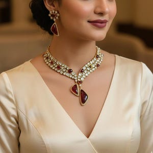 May include: A gold-toned necklace and earring set featuring red and clear gemstones. The necklace has a bib design with a central pendant. The earrings have a matching design. The jewelry is worn with a cream-colored top.