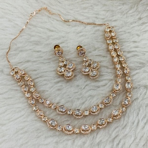 May include: A rose gold jewellery set comprising a double-strand necklace and matching drop earrings. The necklace and earrings are embellished with clear, round gemstones. The set is presented on a white, textured surface.