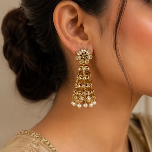 May include: A gold-toned jhumka earring featuring a floral-shaped stud top connected to a multi-tiered, bell-shaped drop. The structure is embellished with clear, round-cut stones set in metal frames and finished with small, white spherical pearls.