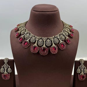 Puede incluir: Un deslumbrante conjunto de collar y pendientes exhibido sobre un busto marrón. El collar presenta un diseño de metal plateado con piedras preciosas claras y rojas. Los pendientes reflejan el diseño del collar, creando un conjunto de joyas cohesivo y elegante.
