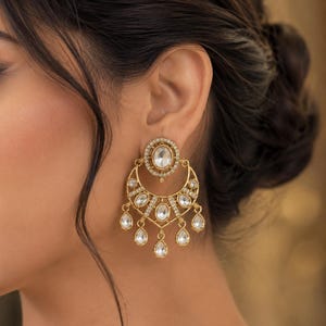 May include: A gold-toned earring featuring a circular stud with a central oval stone, connected to a crescent-shaped drop component. The design is embellished with small clear stones and finished with five dangling teardrop-shaped stones at the base.