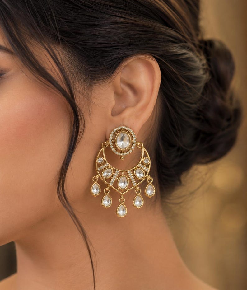 May include: A gold-toned earring featuring a circular stud with a central oval stone, connected to a crescent-shaped drop component. The design is embellished with small clear stones and finished with five dangling teardrop-shaped stones at the base.