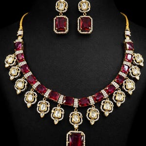 Puede incluir: Un conjunto de collar y pendientes dorados con grandes piedras preciosas rectangulares de color rojo intenso. El collar presenta una serie de piedras rojas y piedras blancas más pequeñas. Los pendientes reflejan el diseño del collar.