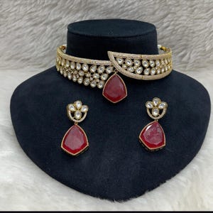 May include: A gold-toned choker necklace and matching earrings set. The choker features a detailed design with clear stones and a large, teardrop-shaped red stone pendant. The earrings have a similar design with red teardrop stones. The jewelry is displayed on a black mannequin head.