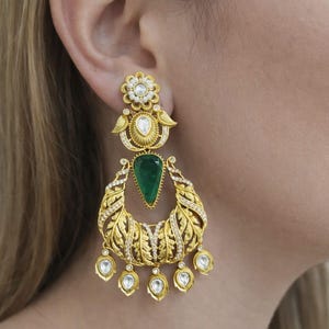 Puede incluir: Pendientes dorados con una parte superior floral, una esmeralda en forma de lágrima y un diseño en forma de media luna con detalles de hojas. Los pendientes presentan piedras claras en forma de lágrima.