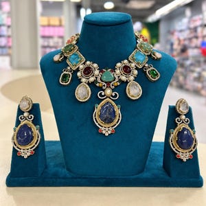 May include: A gold-toned necklace and matching earring set featuring large teardrop-shaped blue stones, accented with smaller multicoloured gemstones, clear crystals, and white pearls, all set in intricate metalwork with a decorative filigree design.