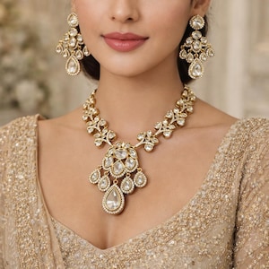 Könnte beinhalten: Ein goldfarbenes Halsketten- und Ohrring-Set. Der Schmuck besteht aus klaren, facettierten Steinen in einem floralen Design. Die Halskette hat einen großen Anhänger mit tropfenförmigen Steinen. Die Ohrringe sind im Chandelier-Stil.