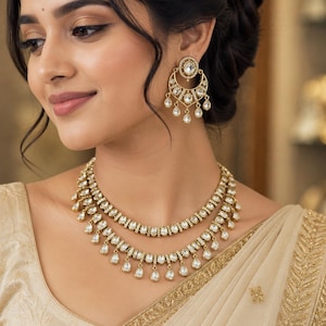 May include: A gold-toned jewelry set consisting of a two-layered necklace and matching crescent-shaped earrings. The pieces feature clear, teardrop-shaped stones set in metal frames, with small dangling accents along the lower edges of the necklace.