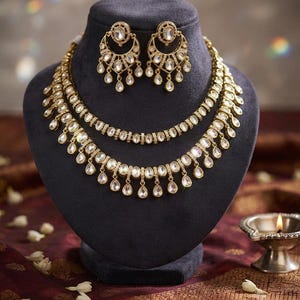May include: A gold-toned jewelry set featuring a two-layered necklace and matching crescent-shaped earrings. Each piece is adorned with clear, oval-cut stones and small, dangling teardrop-shaped pendants, creating a cohesive and ornate appearance.