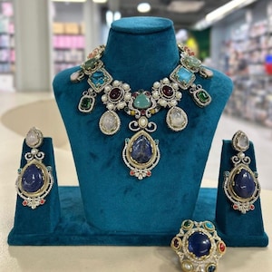 May include: A lavish jewellery set displayed on a teal velvet bust and stands. The set includes a statement necklace, earrings, and a ring. The necklace and earrings feature large, colourful gemstones, pearls, and intricate metalwork. The ring has a large blue gemstone.