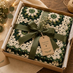 May include: A folded crochet blanket featuring a square pattern in shades of forest green and cream, packaged in a cardboard gift box. The blanket is tied with a dark green satin ribbon and a tag that reads Handmade with love Just for you.