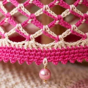 May include: A crocheted lampshade featuring a lattice pattern in vibrant fuchsia and cream yarn. The base of the shade is finished with a solid fuchsia border and a single dangling pink pearl bead accent attached to the bottom edge with a small loop.