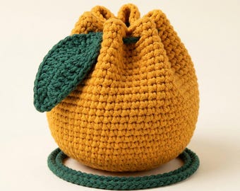 May include: A mustard yellow crochet drawstring pouch shaped like a fruit, featuring a textured stitch pattern, a dark green crocheted leaf attached to the side, and a dark green braided cord strap that encircles the base of the round bag.
