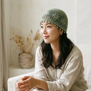 May include: A sage green crochet beanie featuring an intricate openwork floral pattern and a decorative fringe of small matching beads along the bottom edge. The cap is constructed with a fitted crown and a scalloped border detail.