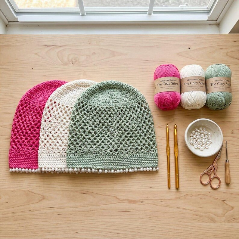 May include: Three crocheted hats in hot pink, cream, and sage green, each featuring a diamond mesh pattern and a white pearl-beaded trim. Included are three matching yarn skeins labelled The Cozy Stitch, two gold crochet hooks, a bowl of pearls, and scissors.