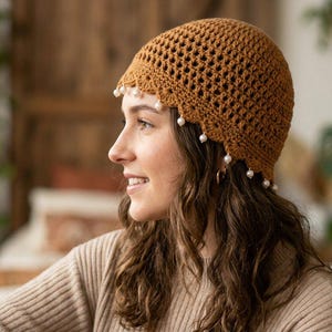 May include: A brown crocheted beanie featuring an openwork lattice pattern throughout the crown and a scalloped edge along the bottom. Small, round, white beads are attached at intervals along the scalloped hem of the hat.