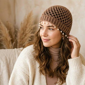 May include: A brown crochet beanie featuring an open diamond-shaped lattice pattern on the crown and a solid ribbed band. The lower edge of the beanie is decorated with small, round, white beads that hang along the forehead and temples.