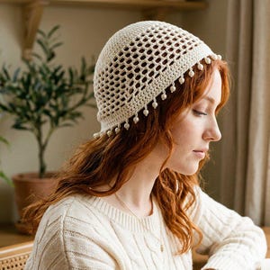May include: A cream-coloured crochet beanie featuring an open-work lattice pattern on the crown and a solid band at the base. The edge of the beanie is decorated with a series of small, dangling cream-coloured beads that hang from the bottom rim.
