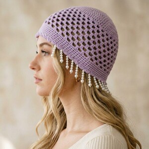 May include: A lavender crochet beanie featuring an openwork diamond pattern throughout the crown. The lower edge of the hat is adorned with a fringe of dangling white faux pearl beads that hang down around the sides and front of the head.