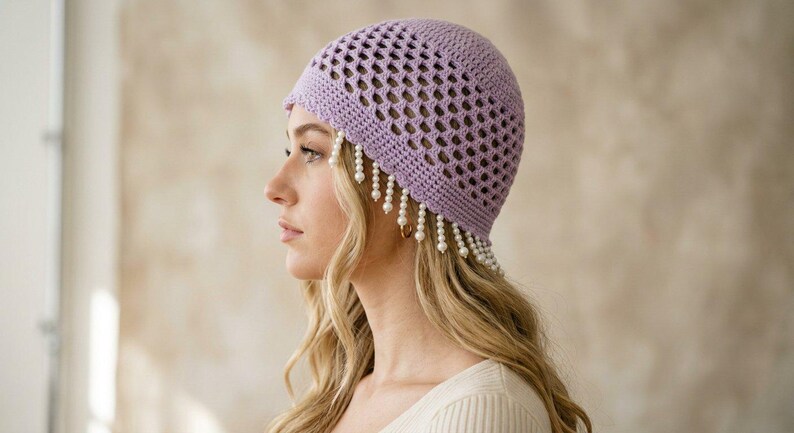 May include: A lavender crochet beanie featuring an openwork diamond pattern throughout the crown. The lower edge of the hat is adorned with a fringe of dangling white faux pearl beads that hang down around the sides and front of the head.
