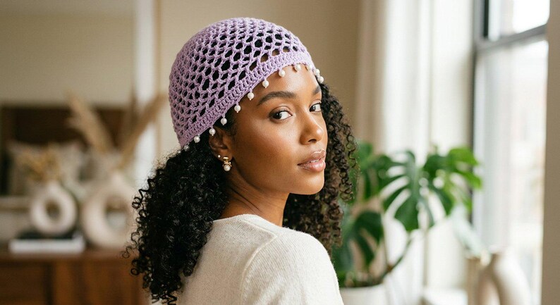 May include: A light purple crochet beanie featuring an open-knit mesh pattern and a decorative border of small white beads dangling along the bottom edge. The hat is constructed from yarn and fits snugly around the crown of the head.