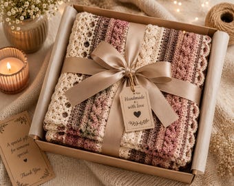 May include: A crocheted blanket with striped patterns in cream, pink, and brown tones, folded inside a cardboard gift box. The blanket is tied with a wide satin ribbon and a tag that reads Handmade with love with a heart icon.