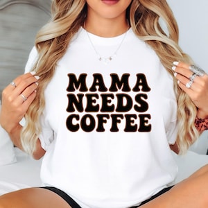 May include: White crew neck t-shirt featuring the text "MAMA NEEDS COFFEE" in black letters with a brown outline. The shirt is a casual style.