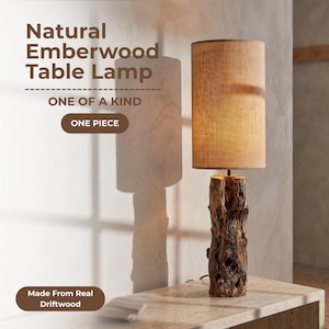 May include: A table lamp with a cylindrical tan fabric shade and a base crafted from a textured, dark brown piece of driftwood. The text on the image reads: Natural Emberwood Table Lamp, ONE OF A KIND, ONE PIECE, Made From Real Driftwood.
