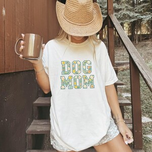 Op de afbeelding: Een cr&egrave;mekleurig t-shirt met de woorden "DOG MOM" in een bloemenpatroon. De persoon op de afbeelding draagt een strooien cowboyhoed en houdt een bruine mok vast. De achtergrond omvat houten trappen en een natuurlijke buitenomgeving.