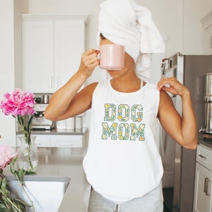 Op de afbeelding: Witte tanktop met de tekst "DOG MOM" in een bloemenontwerp. De letters zijn gevuld met groene bladeren en gele bloemen. De persoon op de foto houdt een roze mok vast en draagt een witte handdoek op het hoofd.