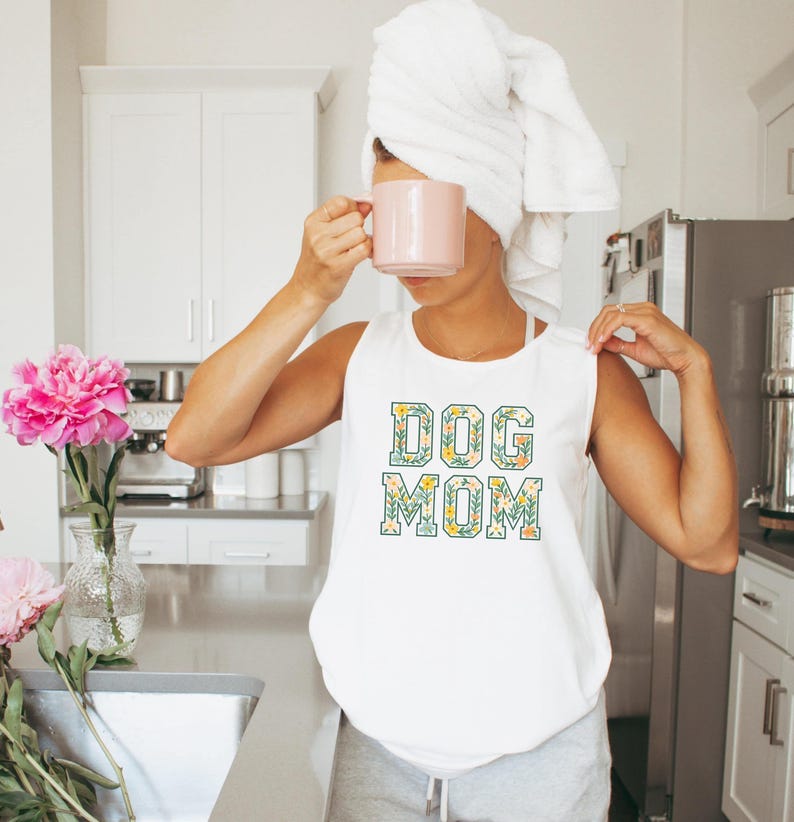 Op de afbeelding: Witte tanktop met de tekst "DOG MOM" in een bloemenontwerp. De letters zijn gevuld met groene bladeren en gele bloemen. De persoon op de foto houdt een roze mok vast en draagt een witte handdoek op het hoofd.