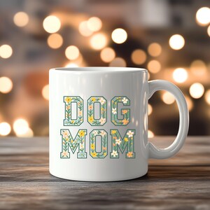 Op de afbeelding: Witte keramische mok met de tekst "DOG MOM" in een bloemenontwerp. De letters zijn groen omlijnd en gevuld met gele, witte en lichtblauwe bloemen en bladeren. De mok heeft een wit handvat.