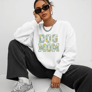 Op de afbeelding: Een witte sweatshirt met de tekst "DOG MOM" in een bloemenontwerp. De sweatshirt wordt gedragen met een zwart-wit gestreepte broek en grijze sneakers. De afbeelding toont een persoon die een zonnebril en een zilveren ketting draagt.