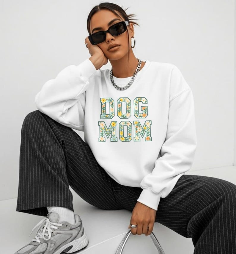 Op de afbeelding: Een witte sweatshirt met de tekst "DOG MOM" in een bloemenontwerp. De sweatshirt wordt gedragen met een zwart-wit gestreepte broek en grijze sneakers. De afbeelding toont een persoon die een zonnebril en een zilveren ketting draagt.