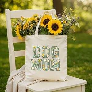 Op de afbeelding: Een cr&egrave;mekleurige draagtas met de woorden "DOG MOM" in een bloemenontwerp. De tas is gevuld met zonnebloemen en andere gele en witte bloemen. De tas staat op een witte stoel.