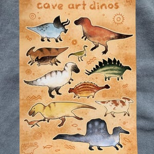 Op de afbeelding: Een rechthoekige print met illustraties van verschillende kleurrijke dinosaurussen op een bruinachtige achtergrond. De tekst "cave art dinos" staat bovenaan, met de naam van de kunstenaar, "Anisaur", onderaan. De dinosaurussen staan in verschillende poses.