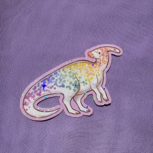 Könnte beinhalten: Ein gestanzter Aufkleber, der einen Parasaurolophus-Dinosaurier mit weißem Körper und einem regenbogenfarbenen, gesprenkelten Muster zeigt. Der Dinosaurier hat einen langen Kamm auf dem Kopf und eine holografische Oberfläche mit Sternreflexen.