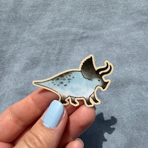 May include: A sticker of a triceratops dinosaur. The dinosaur is light blue with dark blue spots and a dark gray frill and horns. The sticker has a light brown border and is on a light blue background.